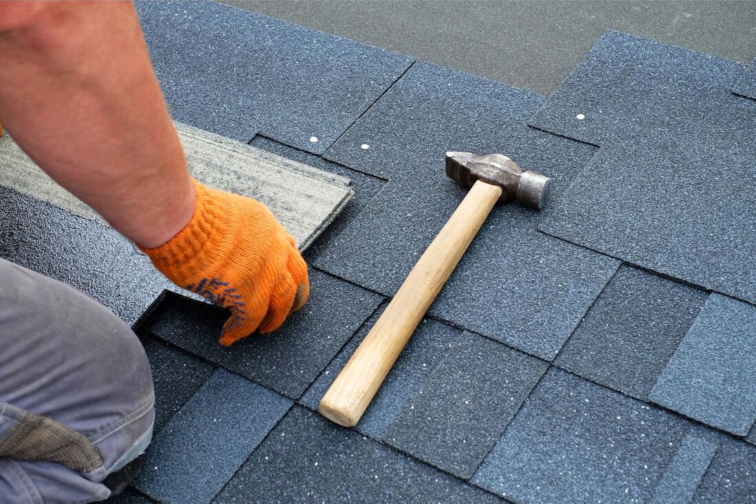 Roofer laying dark grey tiles on a roof. Only his right arm is showing and he is wearing an orange glove while laying a tile. A hammer is placed to the right of the roofer.